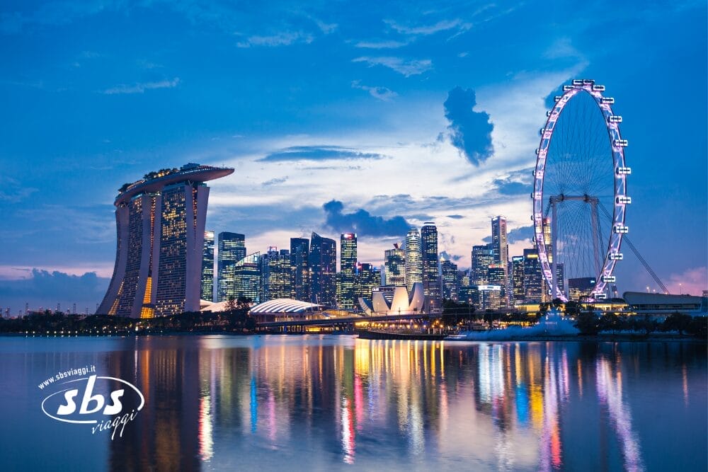Lo skyline di Singapore al tramonto con l'hotel Marina Bay Sands, la ruota panoramica Singapore Flyer e i grattacieli della città riflessi nell'acqua calma sottostante. Perfetto per i viaggi che collegano Singapore a Bali o Gili Trawangan. Il logo "sbs viaggi" compare nell'angolo in basso a sinistra.