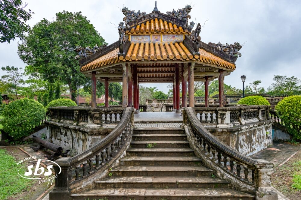 Un padiglione asiatico tradizionale, ornato con un tetto di tegole gialle, intagli dettagliati e scale ricurve, è circondato dal verde e dagli alberi in una giornata nuvolosa, offrendo un'atmosfera tranquilla e automatica alla Bozza.