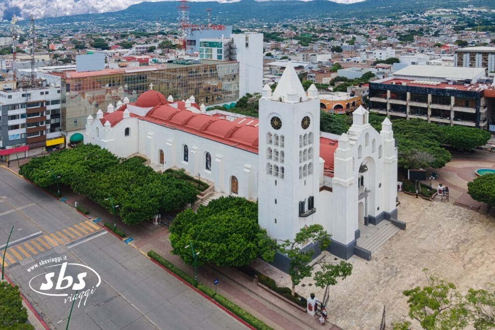 Veduta aerea di una chiesa bianca con un'alta torre dell'orologio, un tetto rosso e finestre ad arco nello Yucatán, circondata da alberi ed edifici cittadini; una grande strada costeggia la chiesa, perfetta per la vostra prossima avventura in Tour Messico.
