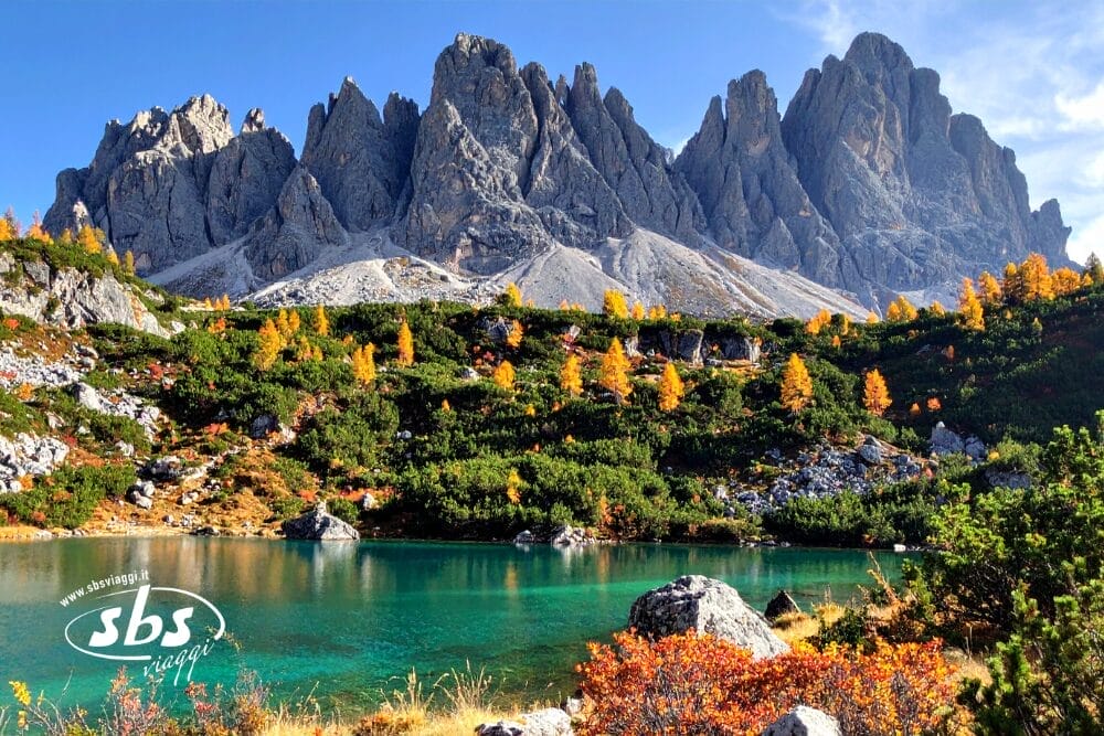 Un lago alpino turchese è circondato da arbusti verdi e alberi autunnali colorati, con le drammatiche montagne grigie e frastagliate che si ergono sullo sfondo sotto un cielo azzurro e limpido, che ricorda i suggestivi paesaggi vicino a Lisbona, nel Sud del Portogallo.