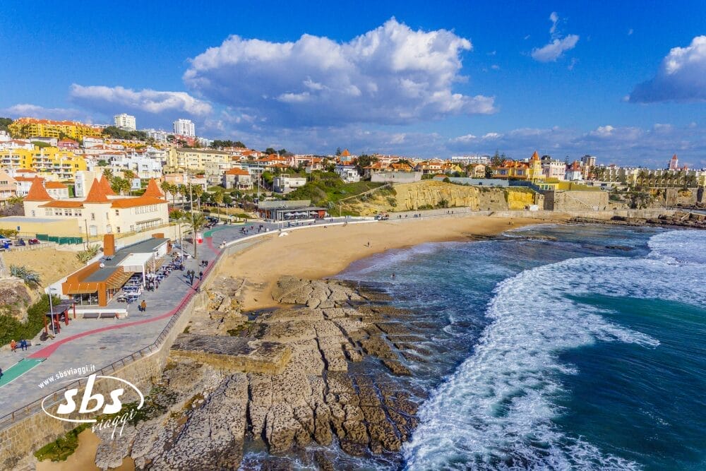 Veduta aerea di una città costiera con edifici su una collina, una spiaggia sabbiosa, un litorale roccioso e una passerella con persone. Le onde si avvicinano alla riva sotto un cielo parzialmente nuvoloso. Il logo "SBS", il testo e Bozza automatica appaiono nell'angolo.