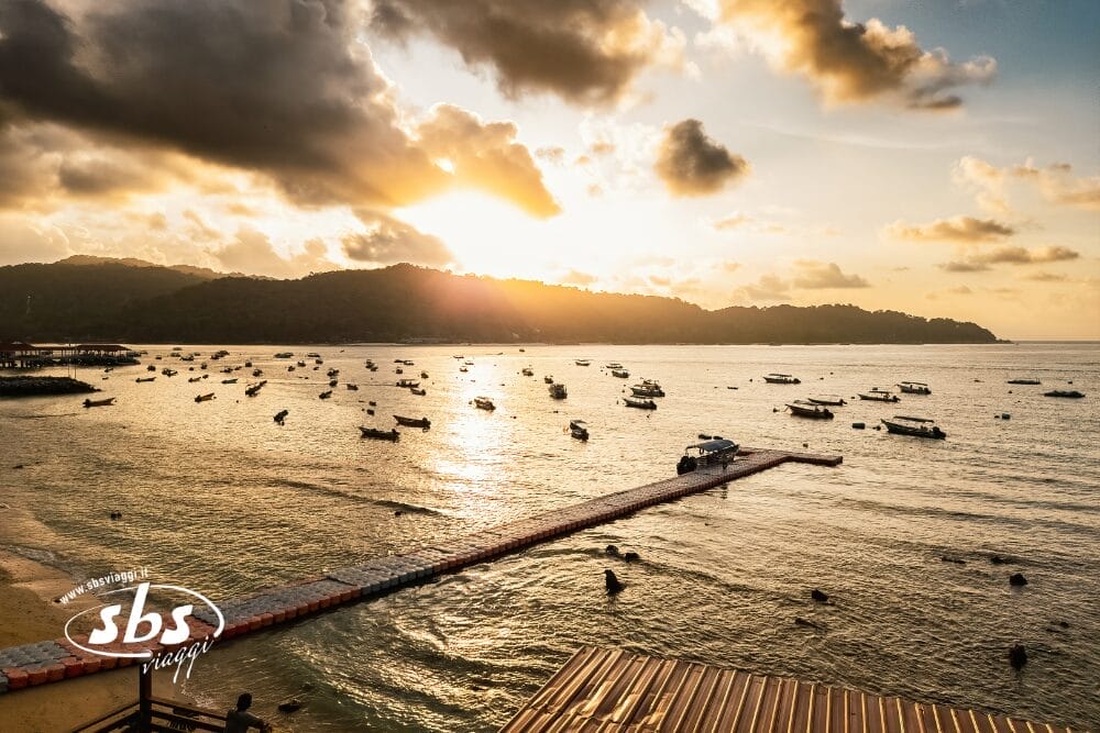 Tramonto su una tranquilla baia delle Isole Perhentian, con molte piccole barche che galleggiano sull'acqua, un molo galleggiante che si protende nel mare e colline sullo sfondo, sotto un cielo parzialmente nuvoloso.
