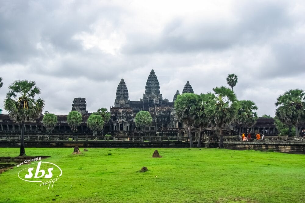 Il tempio di Angkor Wat con le sue iconiche torri sotto un cielo nuvoloso, circondato da palme e erba verde a Siem Reap, con alcune persone vestite con abiti arancioni che camminano vicino al tempio.