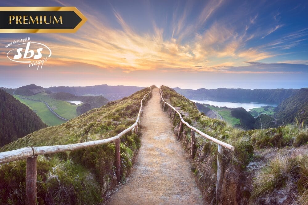 Un sentiero sterrato con ringhiere in legno conduce lungo un crinale di montagna all'alba o al tramonto, con vista su colline verdi e un lago lontano; i loghi "PREMIUM", "sbs viaggi" e "Bozza automatica" appaiono negli angoli superiori.