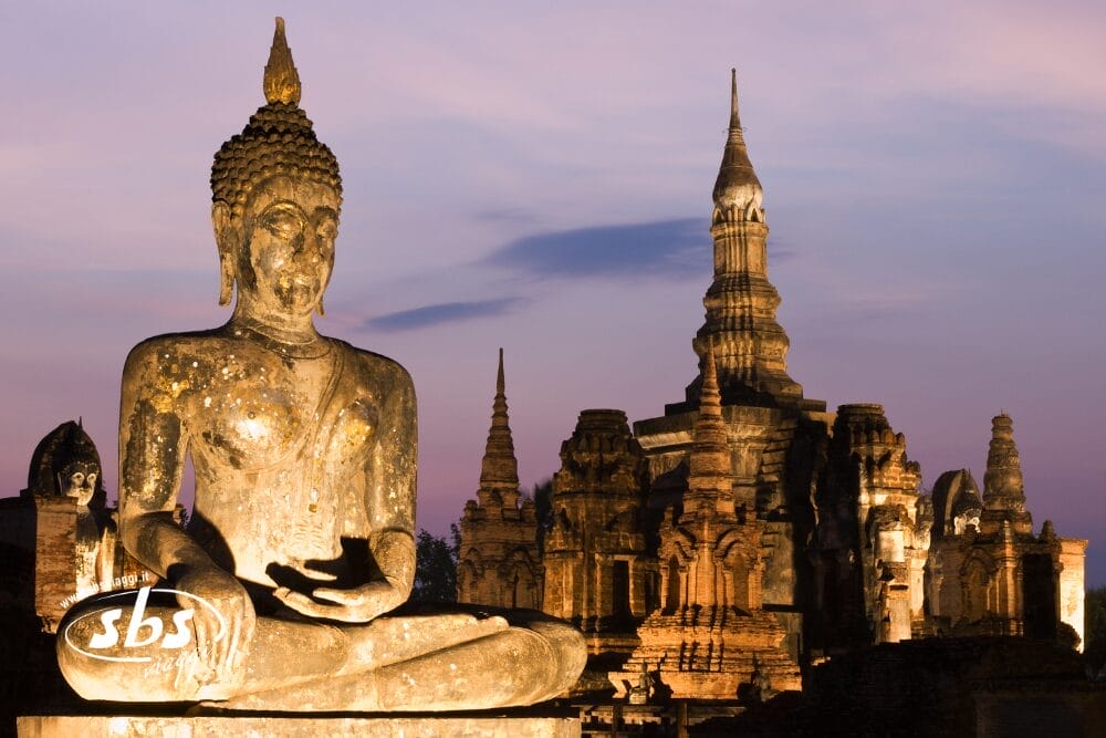 Una grande statua di Buddha in pietra si trova di fronte alle rovine di un antico tempio al tramonto, con morbide sfumature viola e arancioni nel cielo. L'illuminazione calda mette in risalto i dettagli dell'architettura, ricordando una serena Bozza automatica che prende vita.