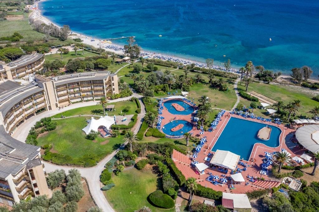 Vista aerea di un resort balneare con edifici curvilinei, grandi piscine all'aperto, lettini, giardini verdi e una spiaggia sabbiosa con acque azzurre e trasparenti: l'ideale per un'esperienza rilassante di Bozza automatica.