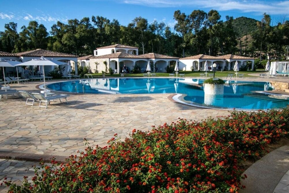 La piscina del resort è circondata da lettini bianchi, ombrelloni ed eleganti ville bianche con tetti di tegole. Fiori rossi e arbusti verdi delimitano l'area della piscina in pietra automatica Bozza, circondata da alberi e colline sotto un cielo blu.