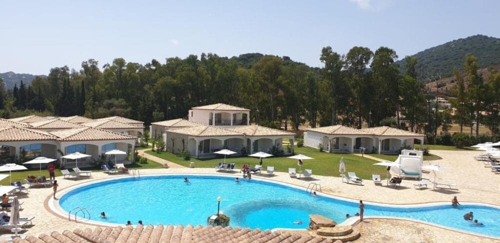 Una grande piscina all'aperto circondata da lettini e ombrelloni, con persone che si rilassano e nuotano. Sullo sfondo, edifici bianchi in stile villa e alberi sotto un cielo azzurro e limpido creano una Bozza automatica perfetta per le vostre vacanze da sogno.