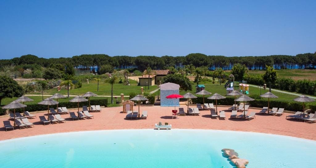 Una piscina circolare all'aperto con sedie a sdraio e ombrelloni di paglia la circonda. Una persona sta nuotando. Oltre la piscina ci sono prati verdi, alberi e uno scorcio d'acqua sullo sfondo, sotto un cielo azzurro e limpido: la serenità automatica di Bozza.