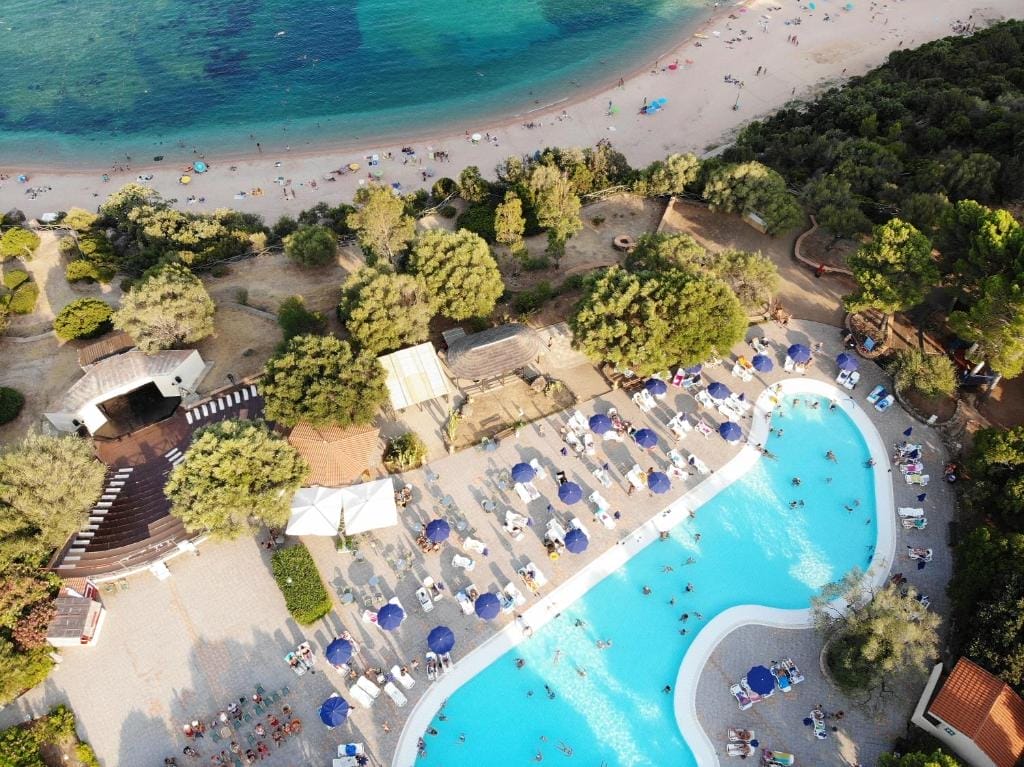 Vista aerea del Club Esse Palmasera Resort, un rifugio 4 stelle a Cala Gonone, con una grande piscina all'aperto circondata da lettini e ombrelloni, accanto a una spiaggia sabbiosa e a un mare cristallino, delimitato da alberi e verde.