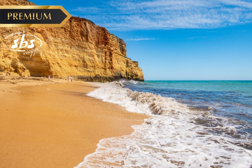 Spiaggia di sabbia dorata con onde dolci e acqua turchese accanto a drammatiche scogliere rocciose sotto un cielo blu brillante. Alcune persone passeggiano lungo il litorale. Nell'angolo in alto a sinistra compaiono i loghi "PREMIUM", "sbs viajes" e la scritta "bozza automatica".