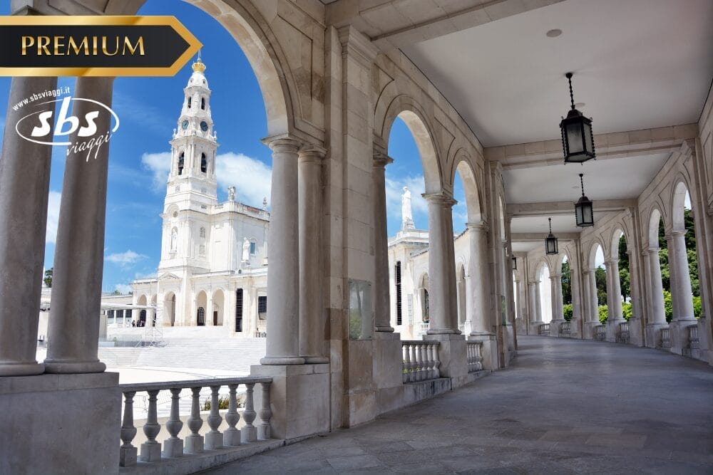 Colonnato di marmo ad arco che si affaccia sulla basilica principale del Santuario di Fátima sotto un cielo azzurro, con i loghi "PREMIUM", "sbs viaggi" e "Bozza automatica" negli angoli superiori dell'immagine.