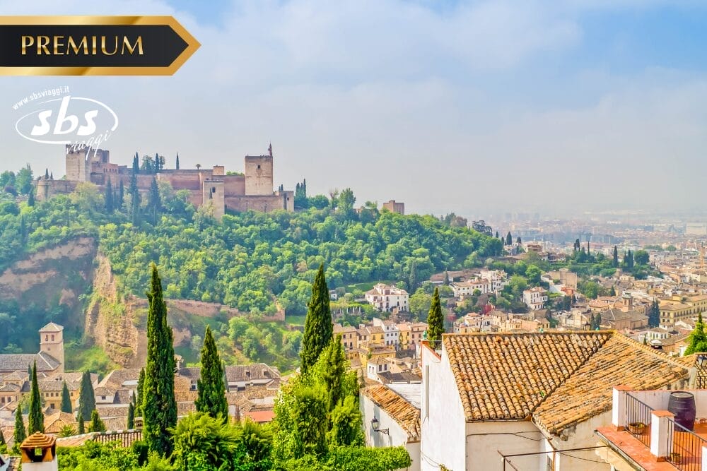 Veduta panoramica di Granada, in Spagna, con lo storico palazzo dell'Alhambra in cima a una collina verde, con gli edifici della città e i tetti di tegole in primo piano sotto un cielo limpido. Sono visibili i loghi "Premium", "sbs" e "Bozza automatica".