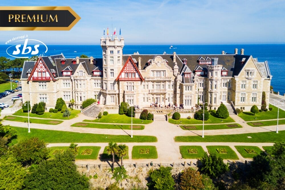 Un grande palazzo storico dall'architettura ornata si trova in riva al mare nella Spagna del Nord, circondato da prati e giardini verdi. I loghi "PREMIUM" e "SBS" sono sovrapposti nell'angolo superiore sinistro dell'immagine.