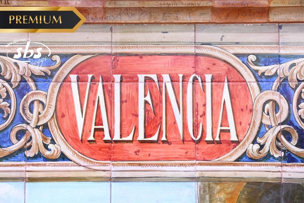 Una piastrella decorativa in ceramica presenta la parola "Valencia" in grassetto a lettere bianche su sfondo rosso, circondata da motivi ornamentali. L'etichetta "Premium" e l'icona di Bozza automatica appaiono in alto, mentre il logo spagnolo di SBS compare in basso.