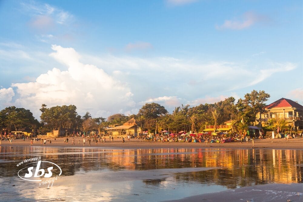Una vivace scena di spiaggia al tramonto con persone che camminano lungo la riva, ombrelloni colorati, alberi ed edifici che si riflettono sulla sabbia bagnata; sopra il cielo parzialmente nuvoloso. Il logo "sbs" e Bozza automatica appaiono nell'angolo in basso a sinistra.