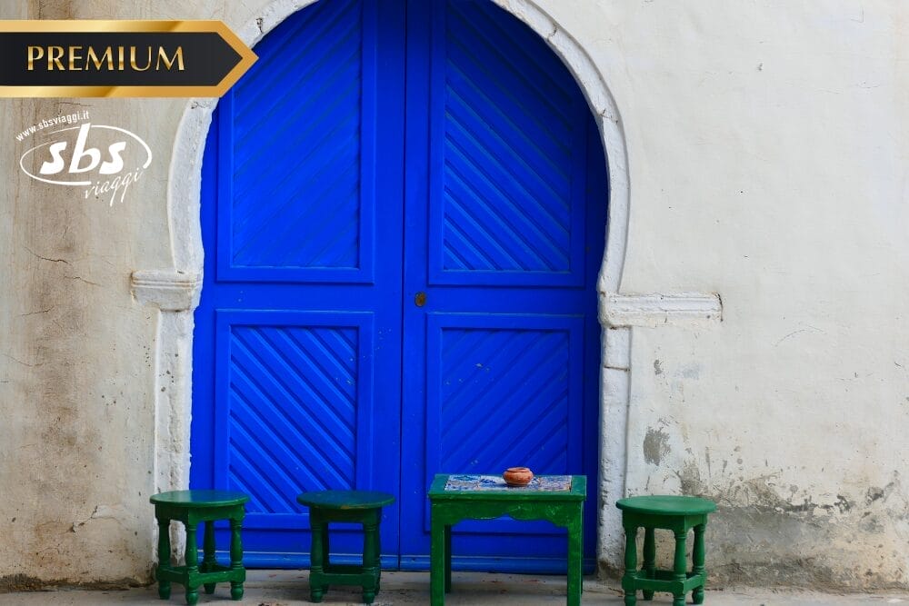 Una porta ad arco blu brillante incastonata in una parete bianca, con tre piccoli sgabelli verdi e un tavolo verde abbinato davanti. Il tavolo contiene una piccola ciotola. Negli angoli dell'immagine compaiono i loghi "Premium", "sbs viaggi" e "Gran Tour Tunisia".