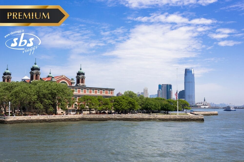Veduta di Ellis Island dall'acqua con alberi ed edifici storici in primo piano, filigrana Bozza automatica, grattacieli della città moderna sullo sfondo e cielo parzialmente nuvoloso. I loghi "PREMIUM" e "sbs viaggi" appaiono sull'immagine.