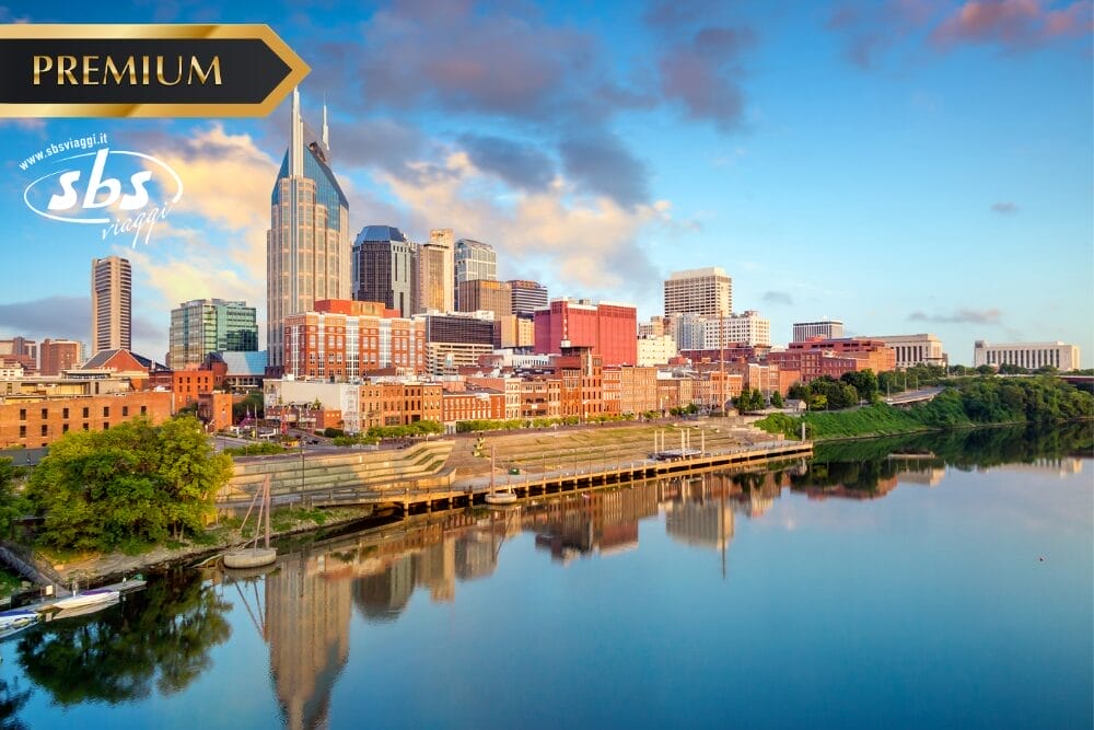 Una vista panoramica del centro di Nashville con edifici moderni, l'iconico edificio AT&T e un fiume tranquillo che riflette lo skyline all'alba, con etichetta "Premium", logo SBS Viaggi e filigrana Bozza automatica nell'angolo.