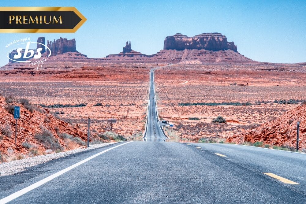 Una strada rettilinea si estende attraverso un paesaggio desertico verso i contrafforti di roccia rossa sotto un cielo blu chiaro, con i loghi "PREMIUM", "sbs" e "Bozza automatica" nell'angolo in alto a sinistra.