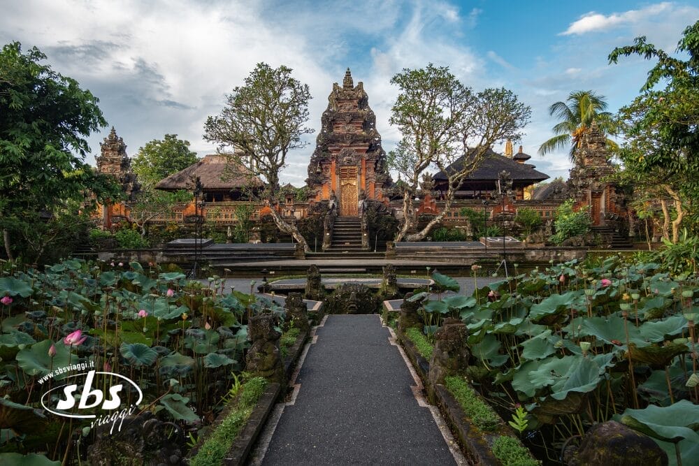 Un sentiero di pietra si snoda attraverso un lussureggiante laghetto di loto fino a un ornato tempio balinese a Ubud, ornato da intricati intagli e circondato da alberi ed edifici tradizionali sotto un cielo azzurro parzialmente nuvoloso.