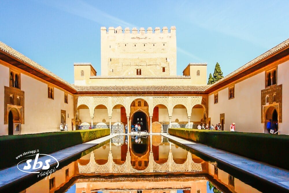 L'immagine mostra la Corte dei Mirti dell'Alhambra a Granada, in Spagna, un punto culminante di qualsiasi Gran Tour dell'Andalusia, con la sua piscina riflettente, le passerelle ad arco, una grande torre e i turisti che passeggiano sotto un cielo azzurro e limpido.