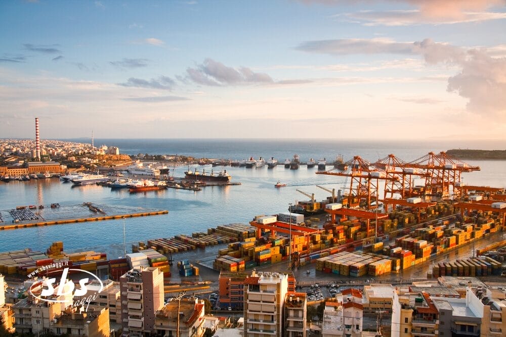 Veduta aerea di un porto affollato che mostra Le Meraviglie del Portogallo, con grandi gru, container e navi attraccate che guidano l'economia sul lungomare, la città e il mare sullo sfondo sotto un cielo parzialmente nuvoloso al tramonto.