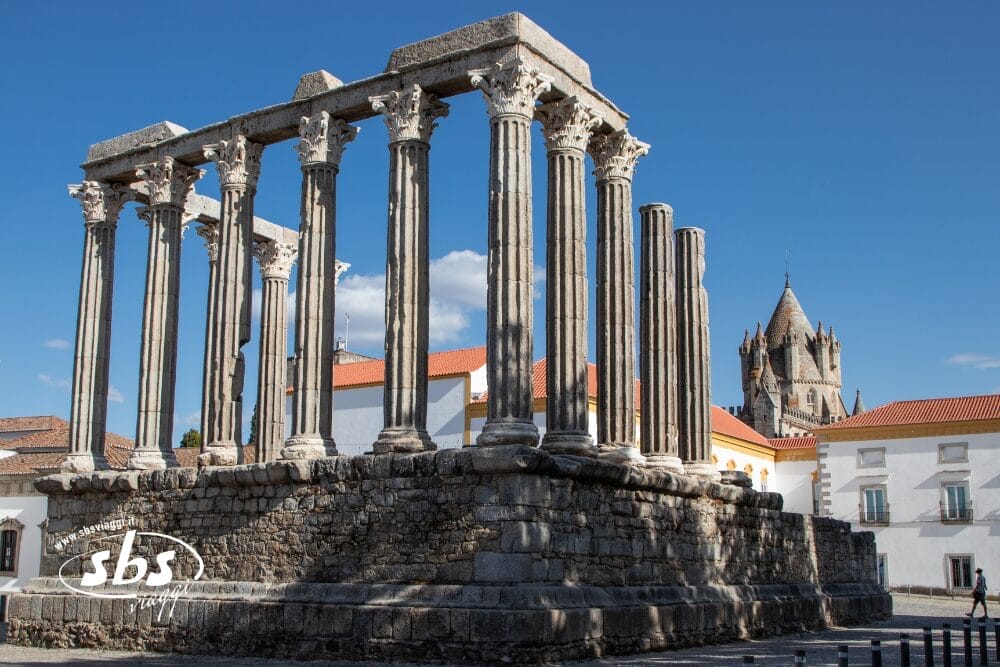 Le rovine di un antico tempio romano con alte colonne di pietra si trovano in una piazza soleggiata nel Sud del Portogallo, circondate da edifici bianchi con tetti rossi; sullo sfondo si erge il campanile di una chiesa sotto un cielo azzurro.