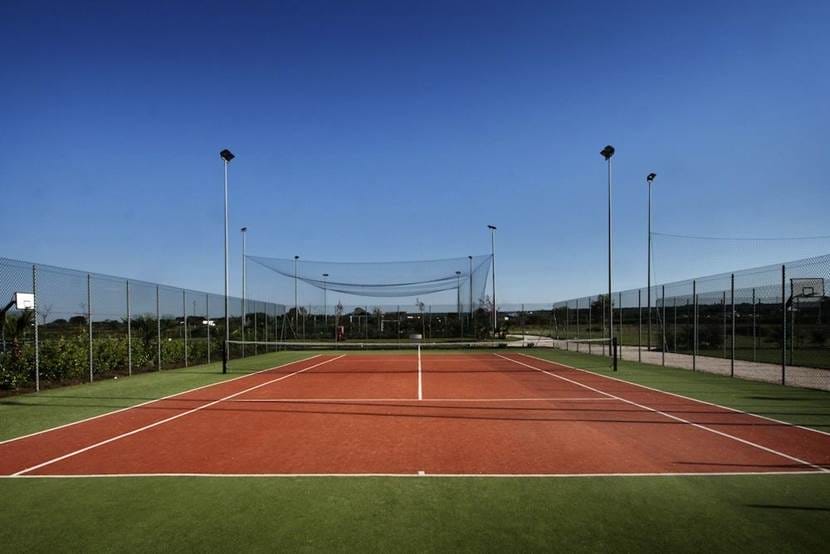 Veduta di un campo da tennis all'aperto vuoto, con superficie di gioco rossa e dintorni verdi, racchiuso da una recinzione di catene, sotto un cielo azzurro e limpido. Bozza automatica: alte luci delimitano il campo e sullo sfondo si intravedono campi aperti.