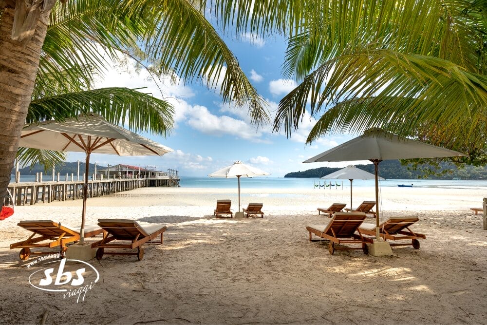 Sedie a sdraio in legno e ombrelloni bianchi costeggiano una spiaggia sabbiosa sotto le palme, di fronte al tranquillo mare blu di Phuket. Un pontile di legno si estende nell'acqua, con colline verdi sullo sfondo: l'ideale per il vostro prossimo Tour Thailandia.