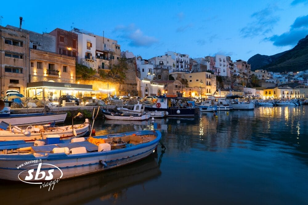 Una vista panoramica di un porto al tramonto vicino a Palermo, con barche da pesca colorate che galleggiano sull'acqua calma ed edifici illuminati che si affacciano sul lungomare sotto un cielo blu intenso. Il logo di SBS Viaggi appare in primo piano e invita a visitare Concordia Sicilia.