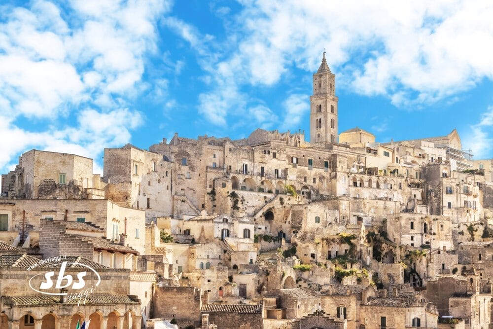 Antichi edifici in pietra e case accatastate sul fianco di una collina sotto un cielo blu brillante con nuvole sparse, con un alto campanile a Matera, Italia. Il logo SBS e "Bozza automatica" appaiono nell'angolo in basso a sinistra.