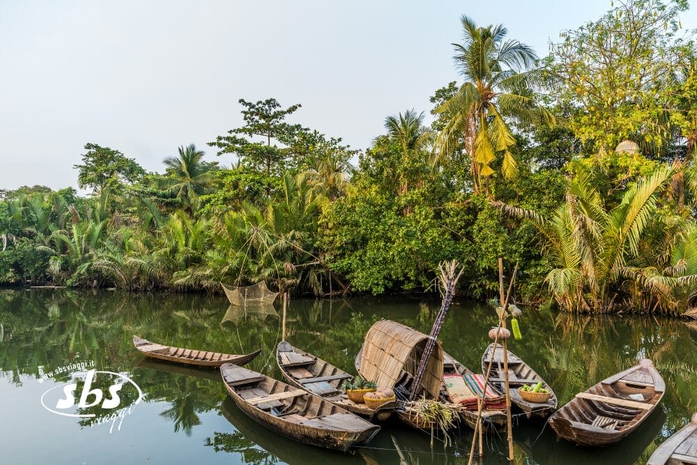 Diverse barche di legno sono ormeggiate su un fiume calmo e verde, circondato da una rigogliosa vegetazione tropicale e da palme, con reti da pesca sullo sfondo: una scena tranquilla che sembra una bozza automatica dell'arte della natura.