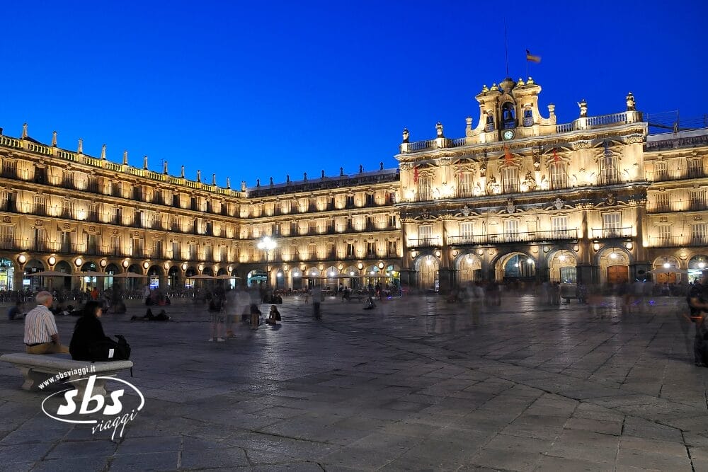 Un grande edificio storico illuminato circonda un'ampia piazza all'imbrunire, con persone sedute e che camminano sulla piazza di pietra. Il cielo è di un blu intenso e la scena ha un'atmosfera vivace e invitante, una vera e propria bozza automatica di vita cittadina.