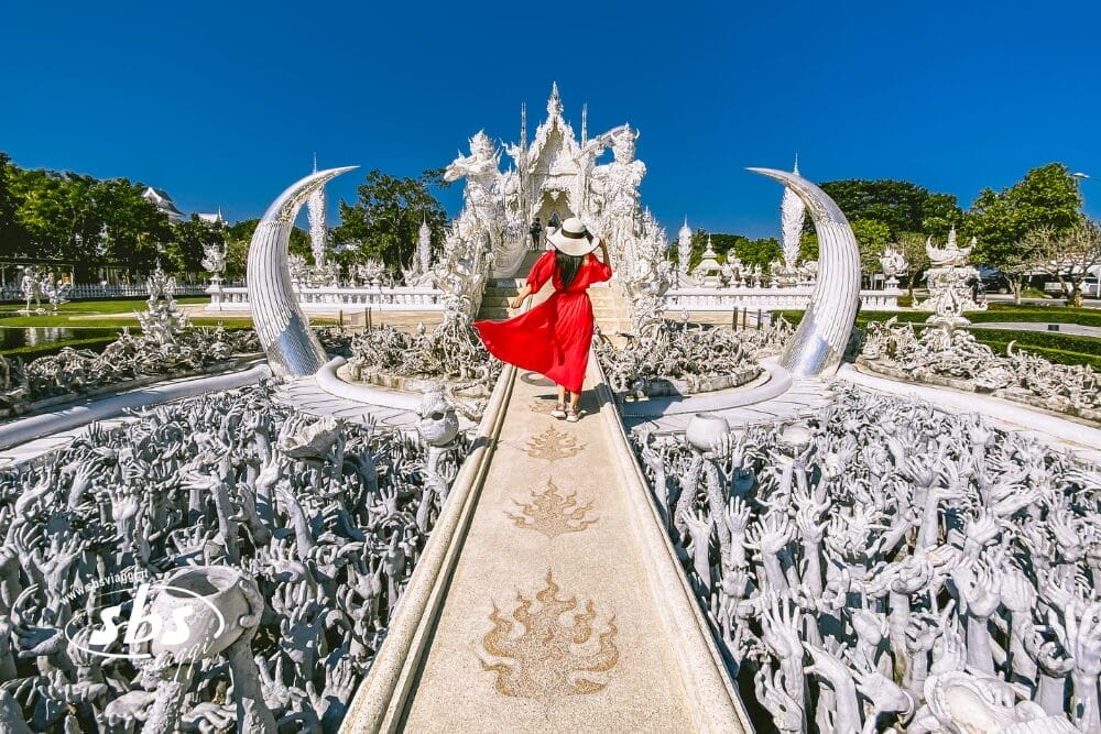Una donna in abito rosso e cappello bianco cammina lungo uno stretto sentiero circondato da mani bianche tese nel Wat Rong Khun, chiamato anche Tempio Bianco, in Thailandia, sotto un cielo azzurro e limpido: una Bozza automatica quasi surreale.