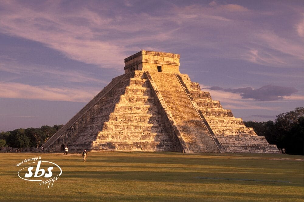 L'immagine mostra l'antica piramide di pietra di Chichen Itza nello Yucatán, in Messico, immersa in una calda luce solare. Il cielo è parzialmente nuvoloso e la struttura sorge su un campo erboso. Il logo "sbs images" è visibile nell'angolo.