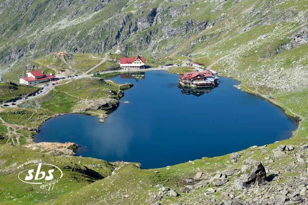 Un limpido lago di montagna circondato da colline verdi e rocciose, con alcuni edifici lungo la riva e una strada tortuosa nelle vicinanze. Il paesaggio, come una bozza automatica, si riflette perfettamente nell'acqua azzurra e calma sotto un cielo luminoso.