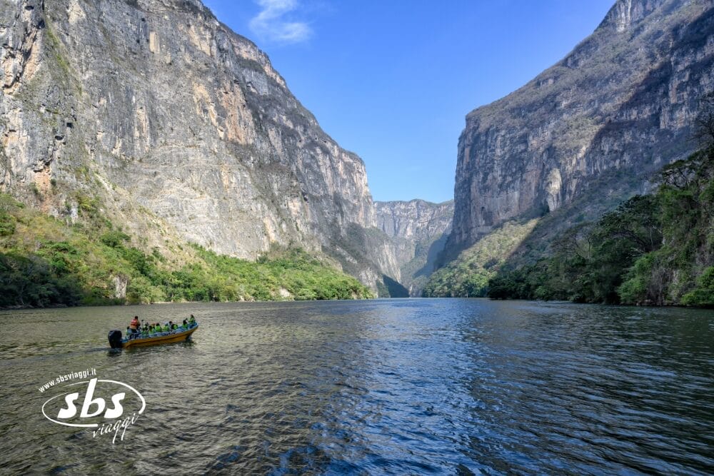 Una piccola imbarcazione con persone naviga su un ampio fiume fiancheggiato da ripide e imponenti scogliere ricoperte di vegetazione, sotto un cielo azzurro con nuvole leggere: una scena iconica del Chiapas, perfetta per un indimenticabile Tour Messico.