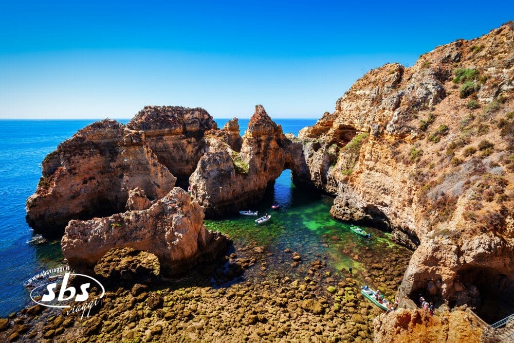 Una scena costiera nell'Algarve, Sud del Portogallo, caratterizzata da scogliere frastagliate e acqua turchese con barche che navigano in archi naturali. Le persone camminano su un sentiero roccioso a destra sotto un cielo azzurro e limpido.