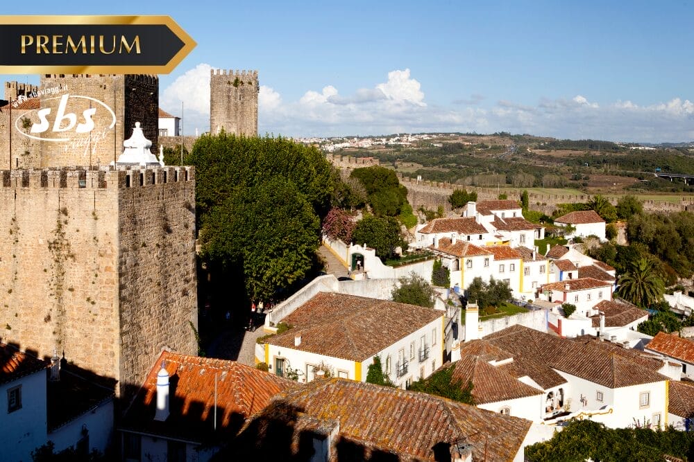 Veduta della torre di un castello storico e delle mura di pietra circondate da case bianche con tetti di tegole rosse in un villaggio illuminato dal sole; "Bozza automatica" appare sotto lo striscione PREMIUM mentre la campagna si estende sotto un cielo blu.