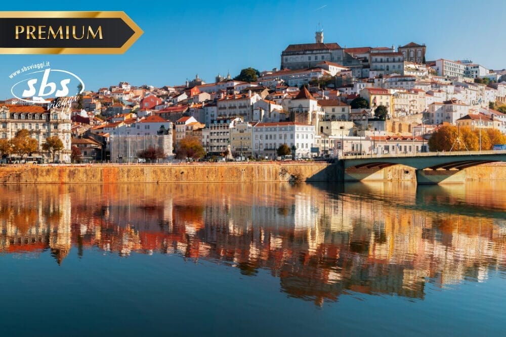 Colorati edifici collinari e una grande struttura storica si affacciano su un fiume con acque calme che riflettono il paesaggio urbano, catturando il fascino del Portogallo Classico. Un ponte attraversa il fiume e un banner "PREMIUM" appare nell'angolo in alto a sinistra.