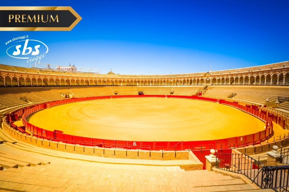 Ampia veduta, illuminata dal sole, di un'arena vuota con sabbia gialla e barriere rosse, circondata da gradinate in pietra sotto un cielo azzurro e limpido. Nell'immagine compaiono i loghi "Premium", "SBS Viaggi" e "Bozza automatica".