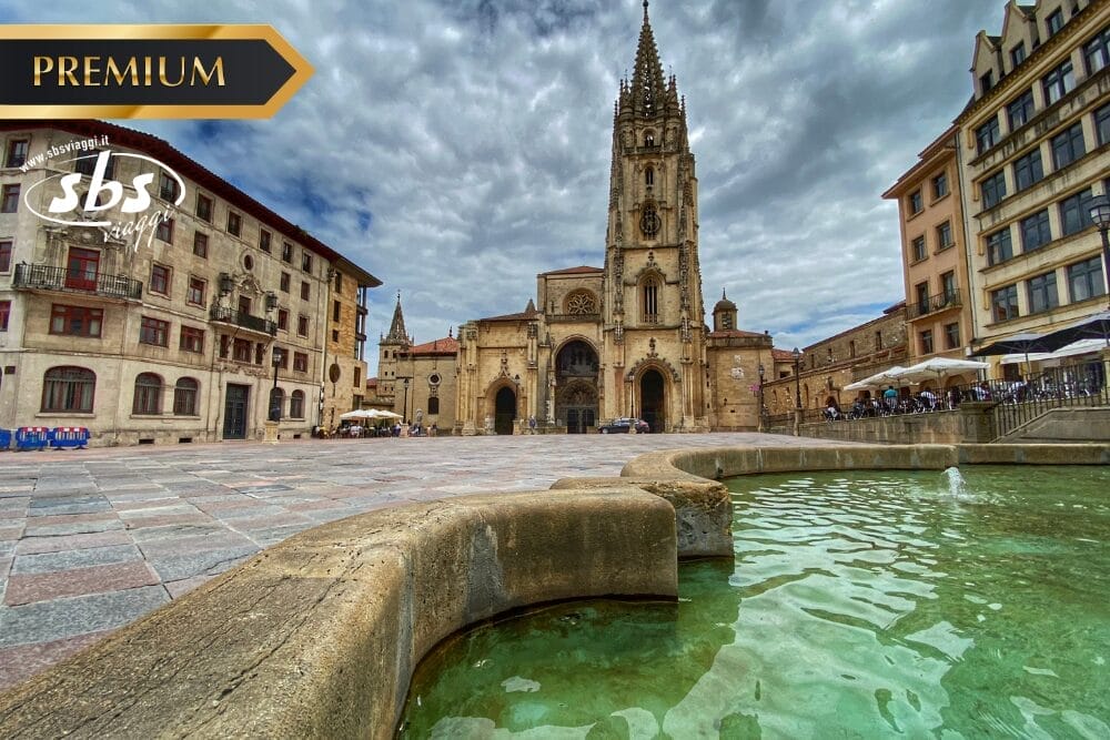 Una piazza in pietra nella Spagna del Nord con una grande chiesa dall'alta guglia sotto un cielo nuvoloso, circondata da edifici storici e da una fontana con acqua verdastra in primo piano.