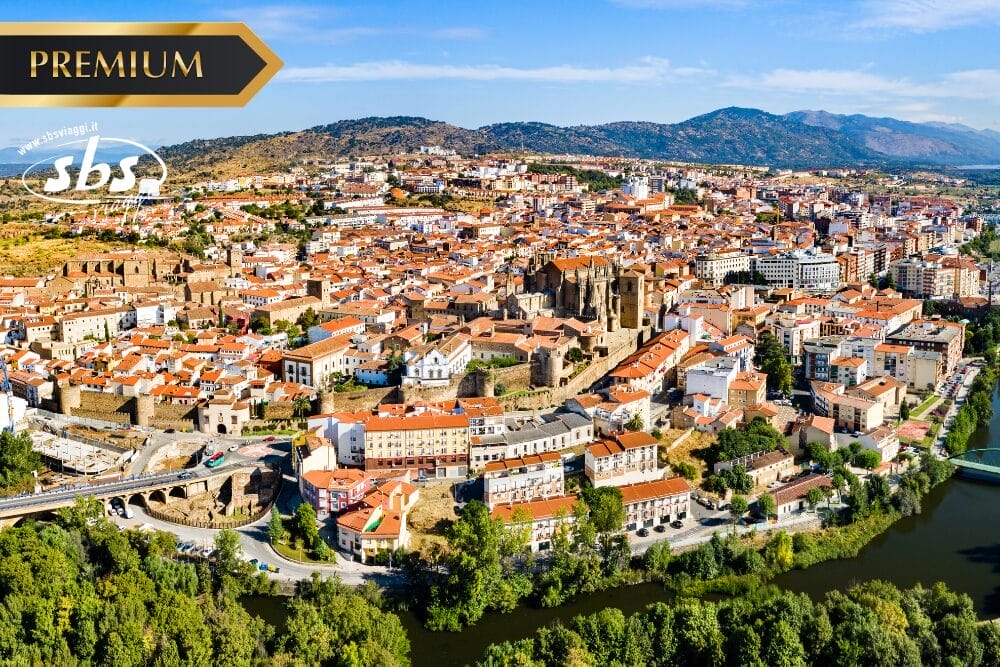Veduta aerea di una storica città spagnola con edifici dai tetti rossi, una cattedrale, strade tortuose e una vegetazione lussureggiante lungo un fiume. L'immagine presenta i loghi "PREMIUM", "sbs" e Bozza automatica per una maggiore autenticità.