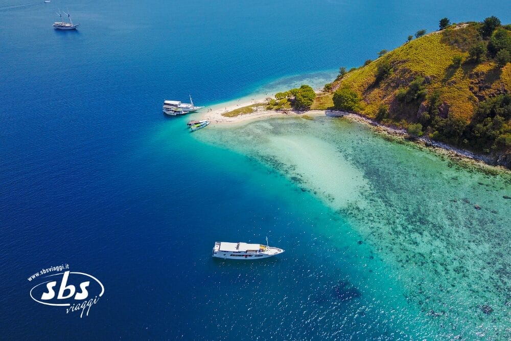 Veduta aerea di una piccola isola lussureggiante con una spiaggia sabbiosa circondata da acque azzurre e trasparenti. Diverse barche sono ancorate vicino alla riva e gli alberi coprono parte dell'isola. Il logo SBS Viaggi e il testo Bozza automatica appaiono nell'angolo in basso a sinistra.