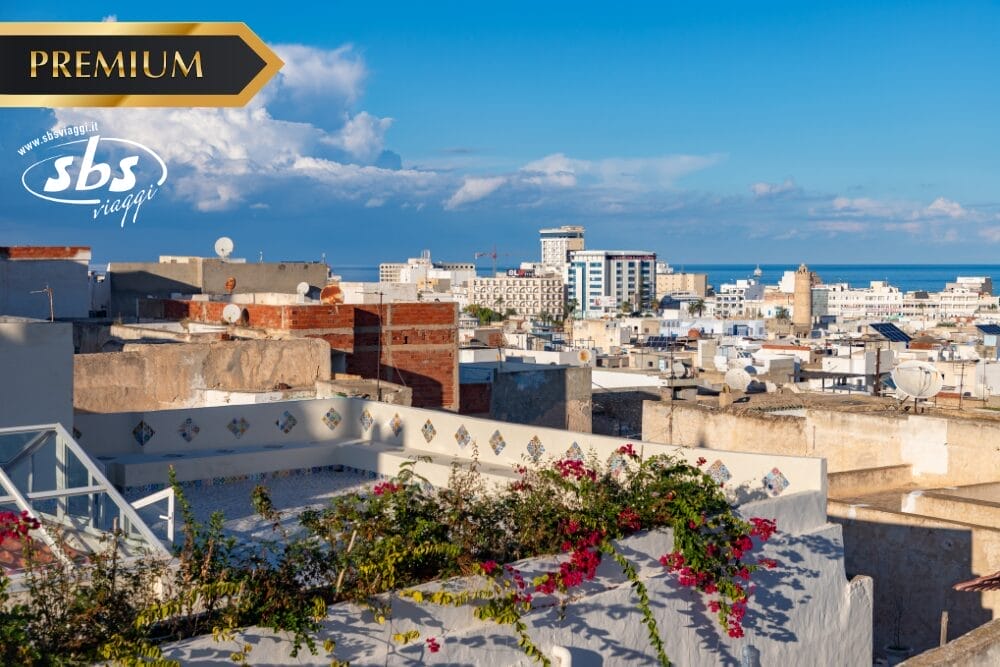 Veduta di una città mediterranea con edifici bianchi, tetti rossi, fiori colorati e una costa lontana sotto un cielo blu. La scritta "Gran Tour Tunisia" e i loghi "PREMIUM" e "SBS viaggi" appaiono negli angoli superiori.