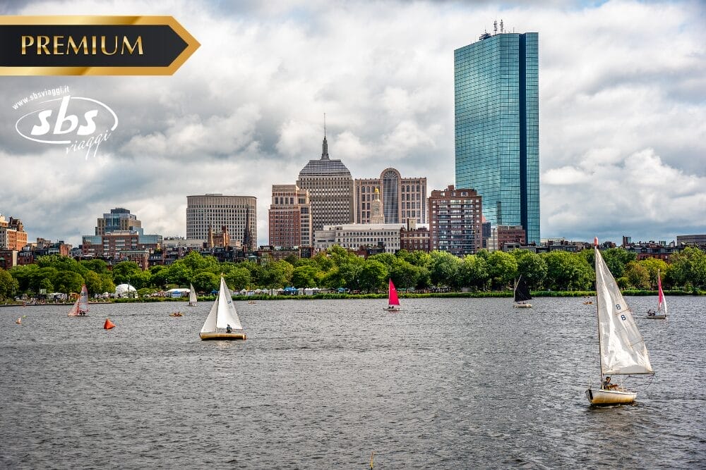 Barche a vela alla deriva su un fiume con lo skyline di New York sullo sfondo, con edifici moderni e storici sotto un cielo nuvoloso. I loghi "PREMIUM" e "sbs" sono sovrapposti all'immagine.
