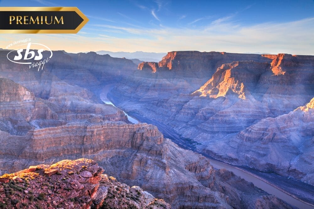 Veduta del Grand Canyon al tramonto, con la luce del sole che illumina le pareti del canyon e un fiume che scorre nella valle. L'immagine include i loghi "PREMIUM" e "sbs viaggi" negli angoli superiori, contrassegnati come Bozza automatica.