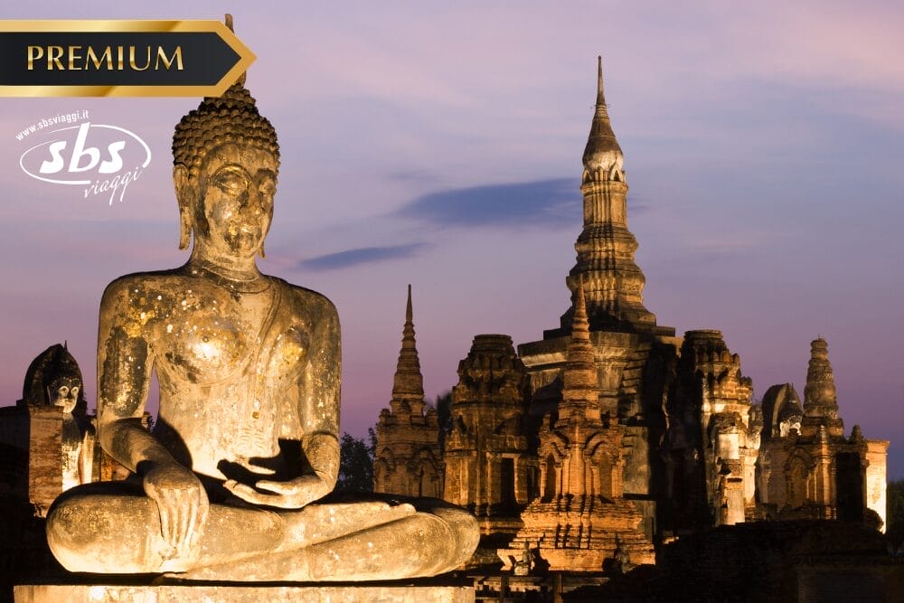 Grande statua antica di Buddha in posizione di meditazione al tramonto, con le rovine e le guglie dei templi storici illuminati sullo sfondo. Scoprite la bellezza spirituale della Thailandia del Nord, da Chiang Mai a Bangkok. I loghi "PREMIUM" e "sbs viaggi" appaiono in alto.
