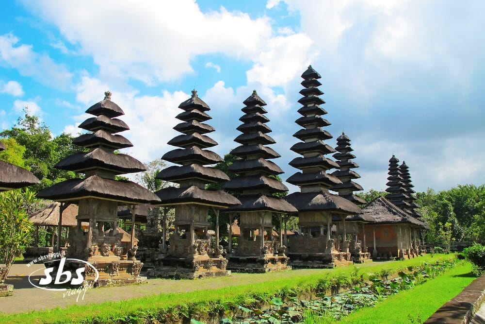 Una fila di templi balinesi tradizionali con tetti di paglia a gradoni si erge tra la vegetazione lussureggiante di Ubud, sotto un cielo parzialmente nuvoloso. In primo piano uno stagno con ninfee e nell'angolo in basso a sinistra il logo bianco "sbs".